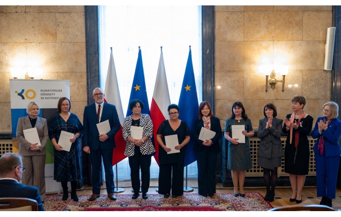 Grupa osób stoi w sali reprezentacyjnej i trzyma dokumenty. W tle widoczne są flagi Polski i Unii Europejskiej oraz baner Kuratorium Oświaty w Katowicach.