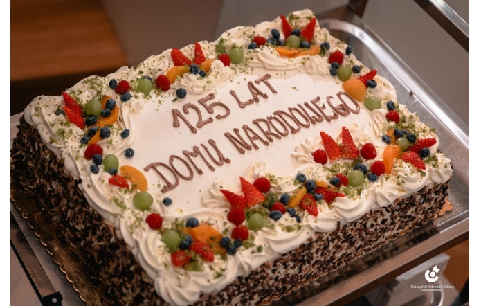 Tort z okazji 125 lecia Domu Narodowego w Cieszynie. Tort prostokątny z kremem, udekorowany owocami i napisem „100 LAT Domu Narodowego”. Na wierzchu widoczne świeczki. 