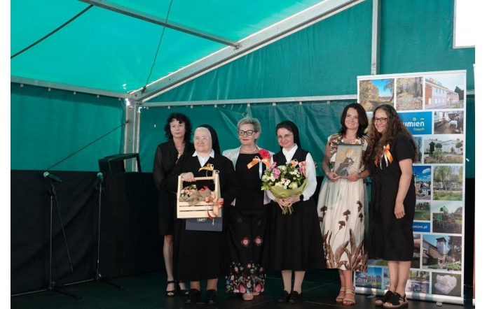 Grupa kobiet pozuje do wspólnego zdjęcia na scenie pod namiotem. Wśród nich są siostry zakonne, jedna z nich trzyma ozdobny upominek, druga bukiet kwiatów, a pozostałe osoby mają przypięte okolicznościowe wstążki.