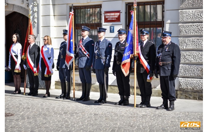 Grupa osób stoi w rzędzie przed budynkiem. Część z nich ubrana jest w mundury strażackie, inne w strój formalny. Kilka osób trzyma sztandary, wszyscy stoją wyprostowani na brukowanej nawierzchni.