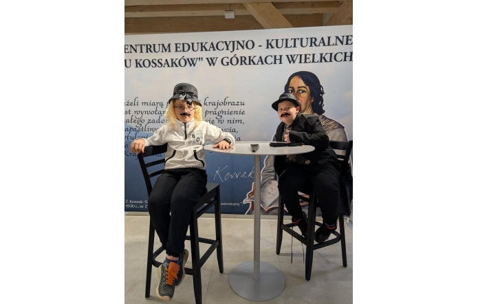 Dwóch młodych chłopców w przebraniach siedzi przy okrągłym stoliku na tle baneru Centrum Edukacyjno-Kulturalnego „U Kossaków” w Górkach Wielkich. Na banerze widać tekst oraz portret kobiety.