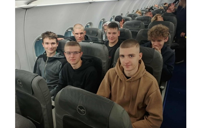 W rzędach szarych foteli samolotu siedzi sześciu nastolatków, większość w ciemnych bluzach; najbliżej po prawej chłopak w brązowej kangurce , obok uśmiechnięci koledzy, a za nimi kolejne rzędy pasażerów