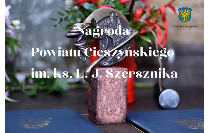 Na pierwszym planie grafiki tekst w kolorze białym – Nagroda Powiatu Cieszyńskiego im. ks. L. J. Szersznika, w tle statuetka wręczana laureatom, składająca się z kamiennego, prostokątnego cokołu oraz mosiężnego odlewu. W prawym górnym rogu herb Powiatu Cieszyńskiego z podpisem.