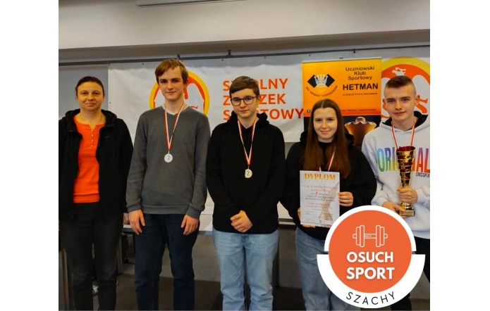 Grupa czterech osób pozuje z medalami, dyplomem i pucharem na tle banerów promujących szachowy turniej sportowy. Trzy młode osoby oraz kobieta w pomarańczowej bluzce stoją uśmiechnięci. W prawym dolnym rogu widoczny symbol z napisem „Osuch Sport – Szachy”.