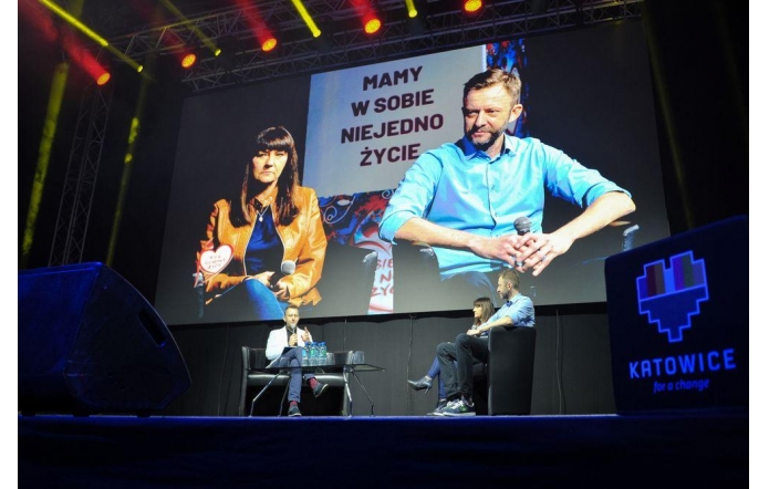 Zdjęcie przedstawia rozmowę na scenie podczas wydarzenia. W tle znajduje się duży ekran, na którym widać dwie osoby: kobietę w brązowej kurtce i mężczyznę w błękitnej koszuli, z mikrofonami w rękach. Na ekranie widnieje napis „Mamy w sobie niejedno życie”. W dolnej części kadru na scenie siedzi trzech rozmówców – mężczyzna w białej marynarce, kobieta i drugi mężczyzna. Na pierwszym planie widać logo miasta Katowice w kształcie serca z kolorowymi paskami i hasłem „Katowice for a change”. Przestrzeń oświetlają jasne reflektory sceniczne.