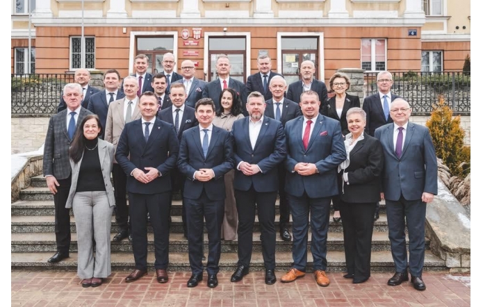 Przed budynkiem na schodach stoi grupa kilkudziesięciu osób ubranych w garnitury i eleganckie stroje. Za nimi widoczna jest elewacja budynku z wejściem i tablicami na ścianie.