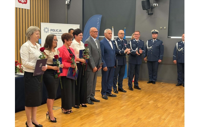 Grupa osób stoi w sali podczas uroczystości. Część osób trzyma róże i teczki, a po prawej stronie stoją policjanci w mundurach. W tle widoczny jest baner Policji oraz godło.