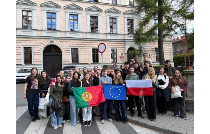Grupa uczniów stoi przed budynkiem i trzyma flagi Portugalii, Unii Europejskiej oraz Polski, pozując do wspólnego zdjęcia.