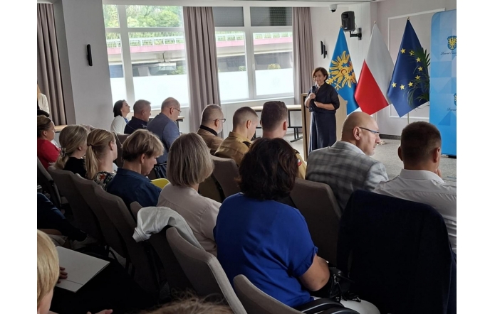 Pełna sala konferencyjna, uczestnicy siedzą w rzędach, zwróceni w stronę mównicy. Przy pulpicie przemawia Janina Żagan Starosta Cieszyński, w tle stoją flagi Polski, Unii Europejskiej i flaga regionalna oraz ścianka z herbem powiatu.