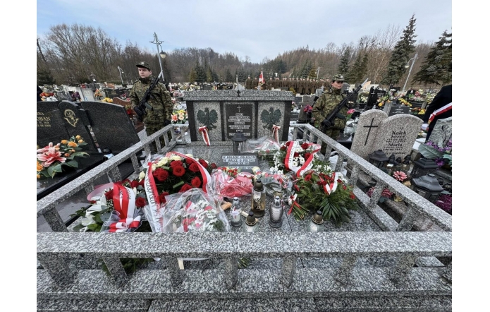 Pomnik z tablicą pamiątkową otoczony wieńcami i zniczami. Wokół zgromadzeni ludzie, część w mundurach. W tle drzewa bez liści oraz zabudowa miejska.