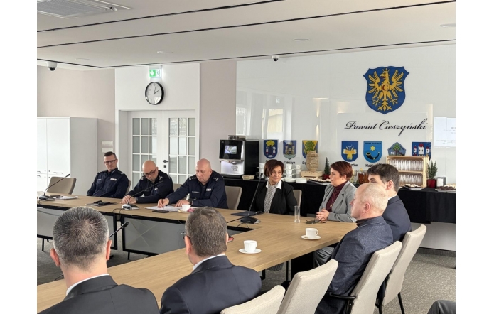 Uczestnicy spotkania siedzą przy stołach ustawionych w podkowę w sali konferencyjnej. Na ścianie widoczny herb, z przodu ekran projekcyjny. Na stołach mikrofony, dokumenty i butelki wody.
