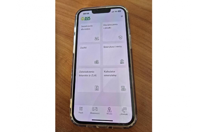 Smartfon z włączoną aplikacją mZUS pokazuje czytelne kafelki z kategoriami takimi jak świadczenia, zasiłki czy emerytury. Leży na drewnianym blacie, w przezroczystym etui, a ekran jest dobrze oświetlony i wyraźny.