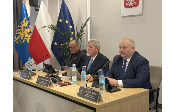 Trzech mężczyzn w garniturach siedzi przy stole konferencyjnym z mikrofonami i tabliczkami. Na stole stoją butelki wody. W tle widoczne flagi Polski i Unii Europejskiej.