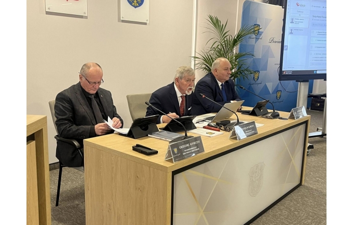 Prezydium Rady Powiatu Grzegorz Konieczny, Stanisław Kubicius, Jarosław Bodak. Na blacie znajdują się dokumenty, tablety, mikrofony i tabliczki imienne, a w tle widać ekran oraz elementy z herbem Powiatu Cieszyńskiego.