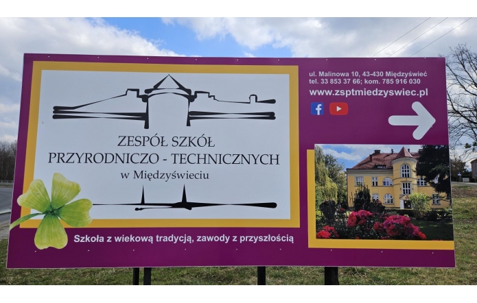 Baner Zespołu Szkół Przyrodniczo-Technicznych w Międzyświeciu ustawiony na zewnątrz. Na fioletowym tle jest nazwa szkoły oraz grafika budynku. Po prawej stronie znajdują się dane adresowe (ul. Malinowa 10, Międzyświeć). Obok umieszczono zdjęcie budynku szkoły otoczonego zielenią i kwiatami. Na dole banera widnieje hasło: „Szkoła z wiekową tradycją, zawody z przyszłością”