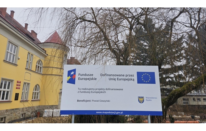 Tablica informacyjna projektu z logotypami instytucji finansujących, ustawiona na zewnątrz, w otoczeniu drzew i zabudowy.