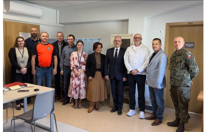 Grupa osób stojących w pomieszczeniu biurowym, ustawionych w rzędzie do wspólnego zdjęcia, wśród nich mężczyzna w mundurze oraz osoby w stroju formalnym i codziennym.