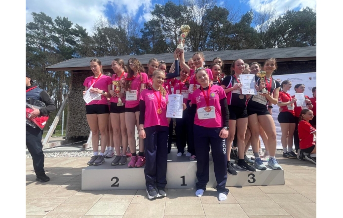 Grupa dziewcząt w różowych koszulkach stoją na podium z pucharami, medalami i dyplomami.