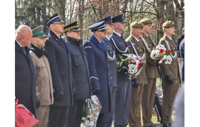 Szereg mężczyzn w mundurach i płaszczach stoi w skupieniu, trzymając wiązanki biało-czerwonych kwiatów podczas uroczystości na świeżym powietrzu.