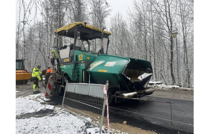 Maszyna drogowa układa asfalt na remontowanej jezdni. Obok pracują osoby w odzieży odblaskowej, a teren robót jest zabezpieczony barierkami. Wokół leży śnieg, a w tle znajdują się drzewa.