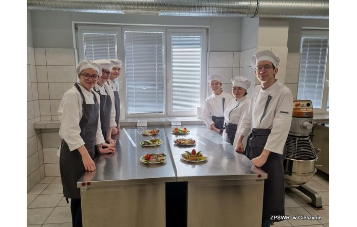 Sześciu młodych kucharzy w białych uniformach i czapkach stoi po obu stronach stalowego blatu, na którym ustawiono talerze z kolorowo podanymi daniami. Tło stanowi jasna kuchnia z oknami i profesjonalnym sprzętem.