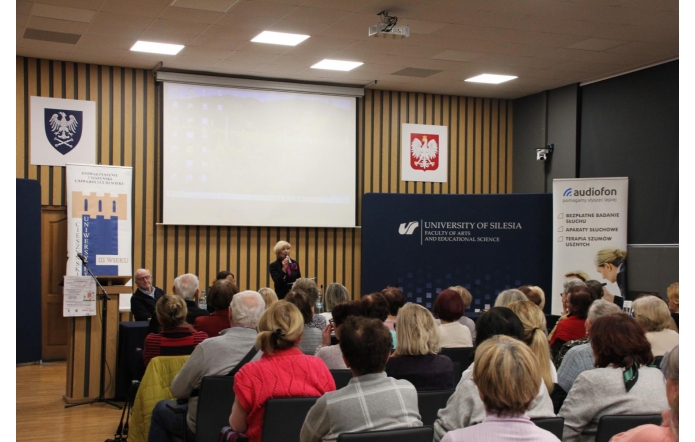 W sali konferencyjnej siedzi liczna grupa osób zwróconych w stronę mównicy i panelu prelegentów. Na scenie stoi kobieta mówiąca do mikrofonu, a za nią widać duży ekran projekcyjny oraz banery informacyjne. Pomieszczenie ma drewniane, pionowe panele na ścianach i emblematy państwowe oraz uczelniane.