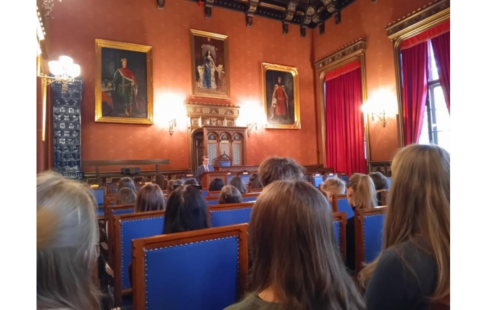 Zdjęcie przedstawia elegancką salę z ozdobnymi, czerwonymi ścianami, w której znajdują się portrety historycznych postaci. W środku stoi osoba przemawiająca do zgromadzonych, siedzących na krzesłach z niebieskimi oparciami. Wnętrze jest bogato zdobione, przy oknach są czerwone zasłony. Wysoki sufit i elementy w stylu klasycznym, podkreślają dostojność pomieszczenia.