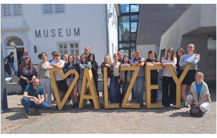 Grupa młodzieży i opiekunów pozuje przed budynkiem muzeum. Uczestnicy stoją wokół dużej przestrzennej instalacji tworzącej napis „ALZEY”, ustawionej na brukowanym placu. W tle widoczna jest jasna elewacja oraz nowoczesna zabudowa z dużymi przeszkleniami.
