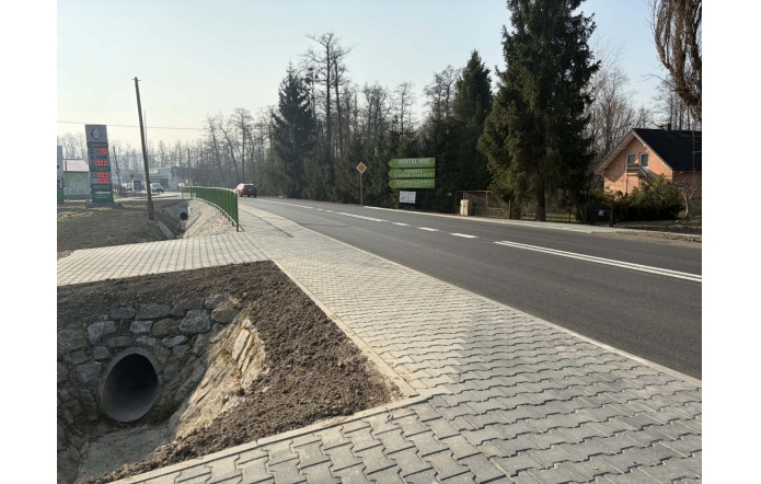 Nowa jezdnia z chodnikiem i barierką przy drodze, obok rów z przepustem, w tle stacja paliw i zabudowania.
