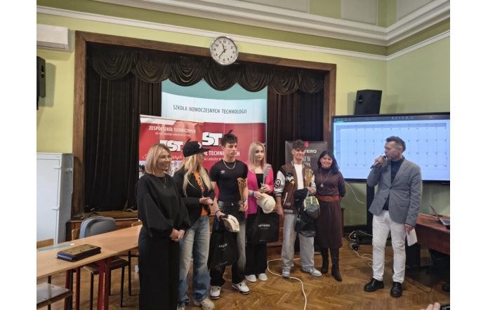 Grupa osób stojących na scenie przed banerem szkoły, część z nich trzyma nagrody i torby, a obok mężczyzna z mikrofonem prowadzi wydarzenie.