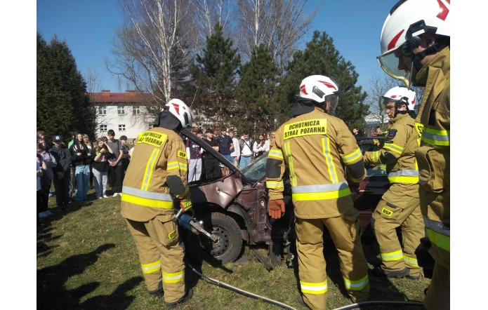 Strażacy z Ochotniczej Straży Pożarnej w pełnym umundurowaniu przeprowadzają pokaz ratownictwa technicznego, używając nożyc hydraulicznych przy uszkodzonym samochodzie. W tle stoi grupa uczniów obserwujących działania.