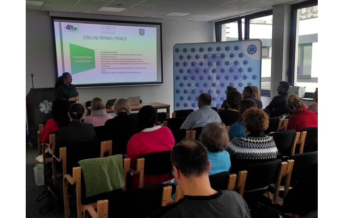 W przestronnej sali konferencyjnej grupa osób siedzi na drewnianych krzesłach, zwrócona w stronę ekranu projekcyjnego. Kobieta w zielonej bluzce prowadzi prezentację dotyczącą usług rynku pracy. Na ekranie widoczne są informacje o poradnictwie zawodowym. W tle duże okna wpuszczają naturalne światło, a na ścianie znajduje się baner z logo ratownictwa medycznego.