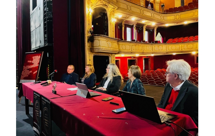 Na scenie zabytkowego teatru przy długim stole z bordowym obrusem siedzi pięć osób z laptopami i mikrofonami, zwrócone w stronę widowni. W tle rzędy czerwonych foteli i złocone balkony, po lewej na sztaludze plakat festiwalowy.