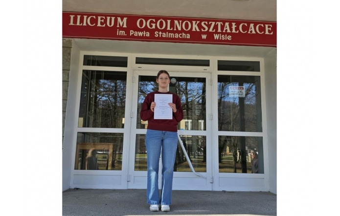 Dziewczyna stoi przed wejściem do liceum i trzyma w rękach dyplom, nad drzwiami widnieje napis z nazwą szkoły w Wiśle.