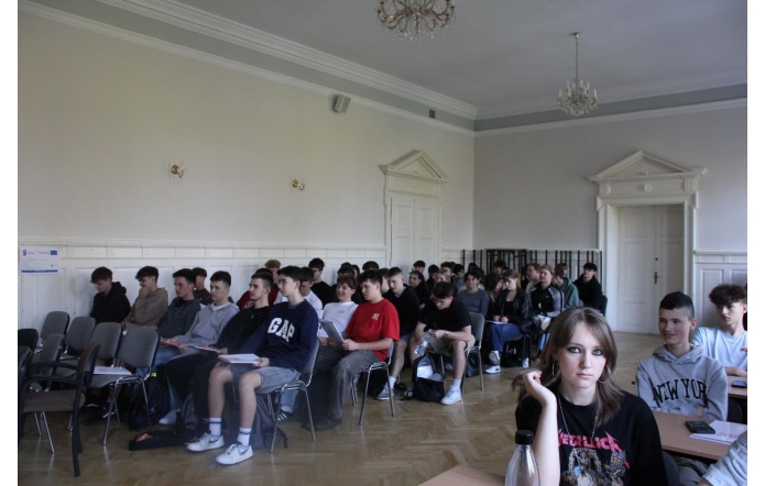 Sala szkoleniowa z grupą uczniów uczestniczących w kursie w ramach projektu unijnego. Młodzież siedzi na krzesłach, trzymając materiały edukacyjne. W tle widoczne drzwi, białe ściany i tablica informacyjna o dofinansowaniu UE.