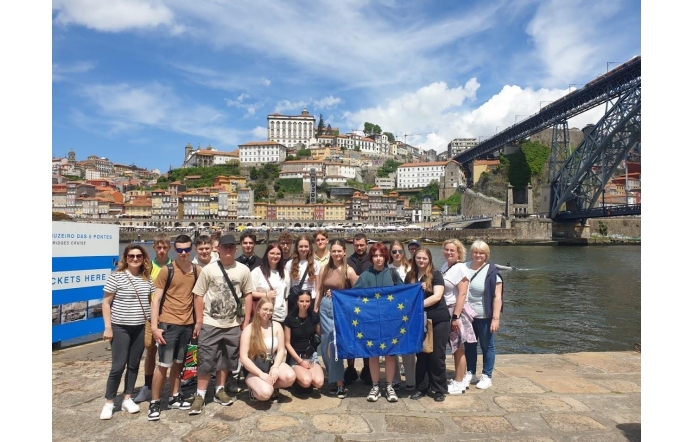 Grupa młodzieży i kilka dorosłych osób pozujących nad rzeką w Portugalii. W tle widać kolorowe kamienice i most. Jedna z dziewczyn trzyma flagę Unii Europejskiej. Zdjęcie zrobiono w słoneczny dzień.