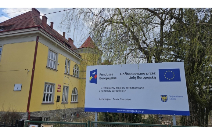 Przed żółtym budynkiem szkoły stoi duża tablica z informacją o dofinansowaniu z Funduszy Europejskich, obok widać drzewo i ogrodzenie.