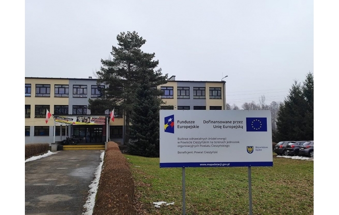 Budynek szkoły widoczny za drzewami i chodnikiem. Na trawniku przed budynkiem stoi tablica informująca o projekcie dofinansowanym przez Unię Europejską.