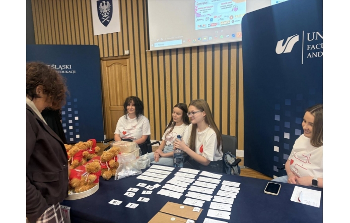 Kilka osób siedzi przy stole rejestracyjnym w sali konferencyjnej. Na stole leżą kartki z numerami, telefon, pojemniki oraz pluszowe misie. W tle widoczne są banery uczelni.