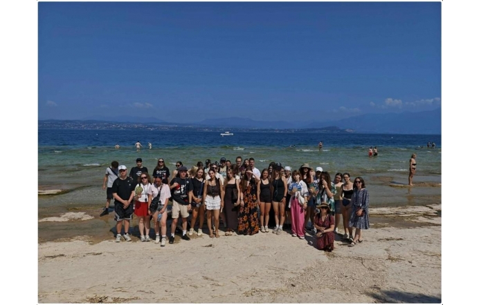 Grupa osób pozuje do zdjęcia na plaży. W tle widać wodę, kąpiących się ludzi, odległy brzeg oraz góry pod błękitnym niebem.