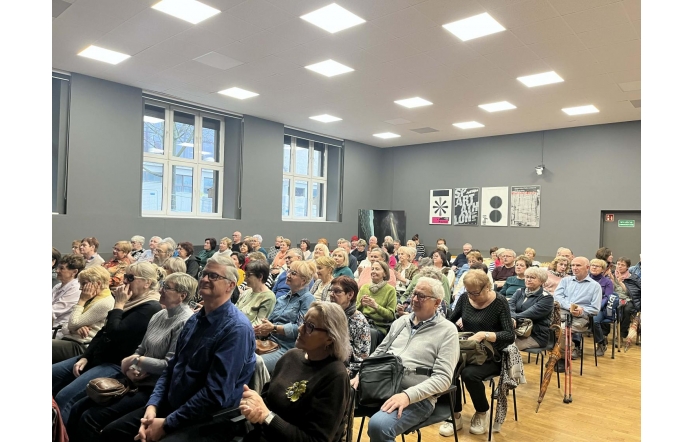 Duża grupa osób siedzi na krzesłach, zwrócona w jedną stronę, część trzyma w rękach torby lub ubrania, a niektórzy mają obok siebie laski.