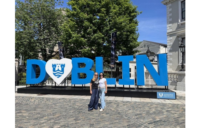 Dwie kobiety pozują do zdjęcia przy dużym napisie „Dublin” w niebiesko-białej kolorystyce. W tle widać drzewa, zabudowę miejską oraz słoneczne niebo.