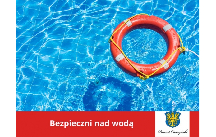 Jasnoniebieska woda basenu z delikatnymi refleksami słońca, po której unosi się pomarańczowe koło ratunkowe z białymi odblaskami i żółtą liną. W dolnej części grafiki czerwony pasek z napisem „Bezpieczni nad wodą” oraz herbem i logotypem Powiatu Cieszyńskiego.