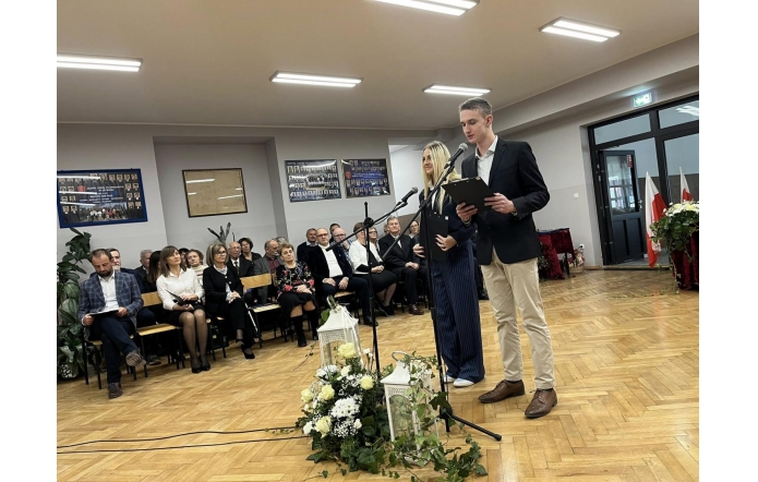 Dwie osoby stoją przy mikrofonach w sali szkolnej. W tle widać siedzącą publiczność, dekoracje kwiatowe oraz flagi.