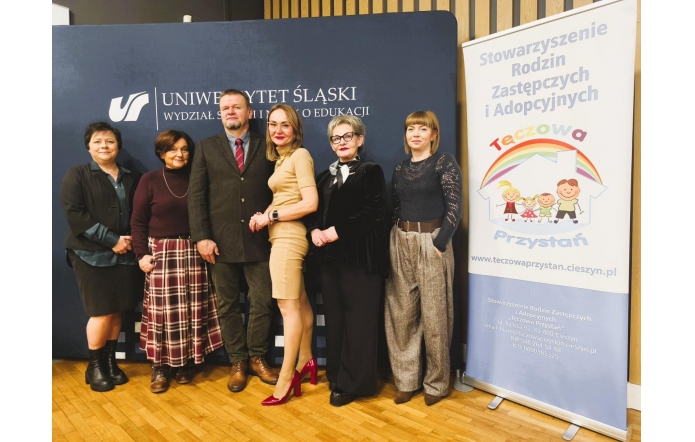 Grupa osób pozuje do zdjęcia na tle baneru Uniwersytetu Śląskiego oraz roll-upu Stowarzyszenia Rodzin Zastępczych i Adopcyjnych „Tęczowa Przystań”.